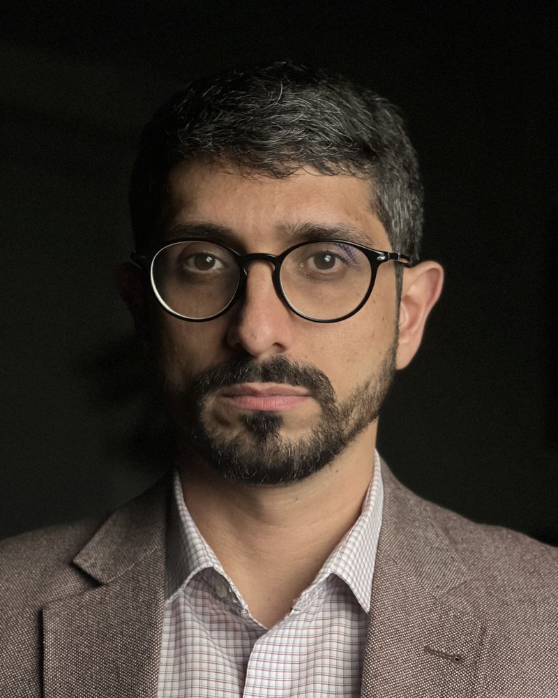 Retrato de Dario Leitão, de óculos e blazer, sobre fundo escuro